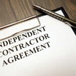 Clipboard with independent contractor agreement and pen on wooden desk background